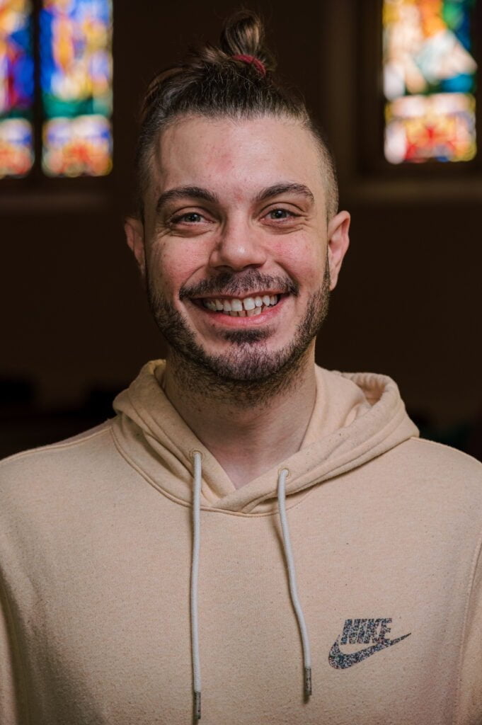 A portrait of Gabe Adorni, church school / nursery assistant, standing in front of the stained glass windows in the sanctuary