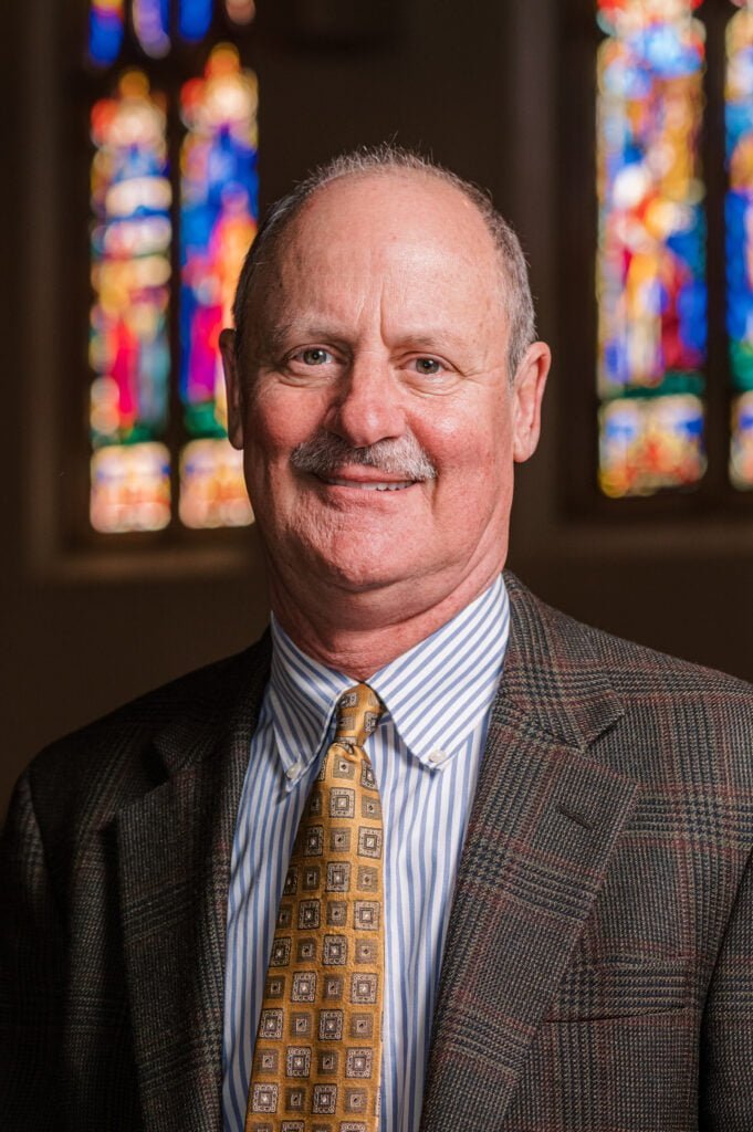 A portrait of Ken Baily, senior pastor, , standing in front of the stained glass windows in the sanctuary