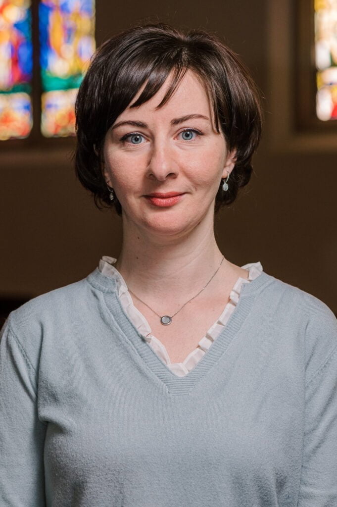 A portrait of Yevgenia Semeina, organist, standing in front of the stained glass windows in the sanctuary