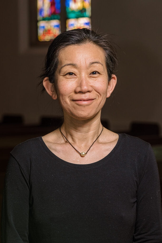 A portrait of Yoko Bryden, building administrator, standing in front of the stained glass windows in the sanctuary