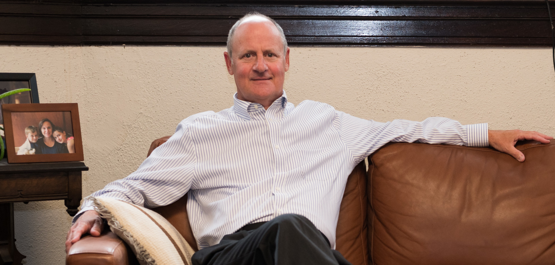 A smiling Rev. Dr. Ken Baily sitting comfortably on a brown leather couch with his arm extended, wearing a striped button-up shirt and dark pants, with a framed photo on a side table beside him.